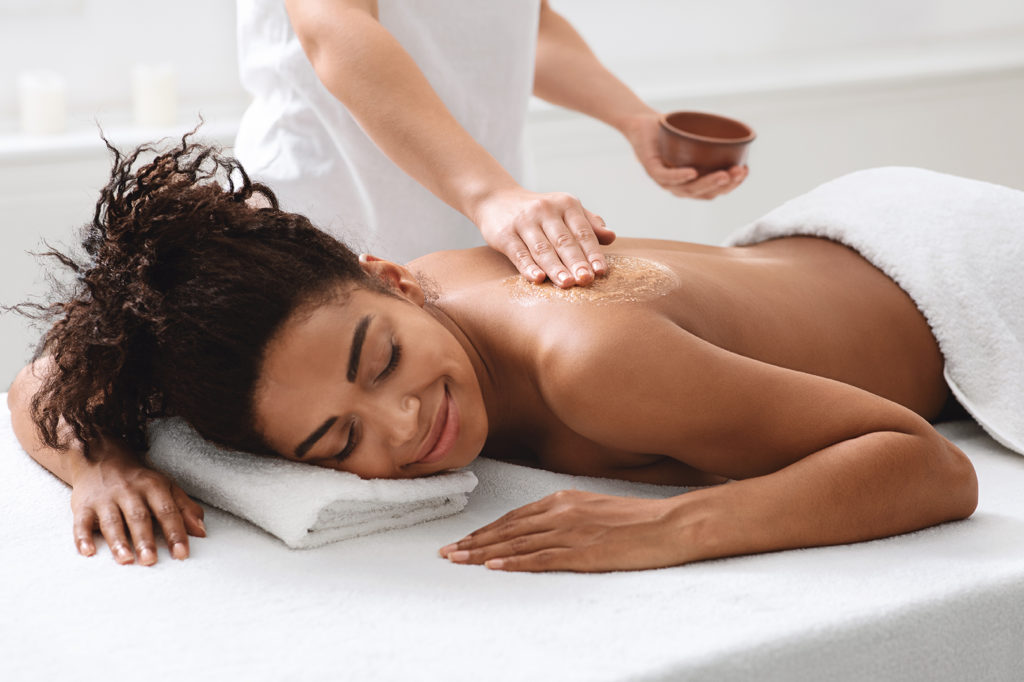 Spa therapist applying exfoliating body mask on sleeping black woman back at luxury spa, body care concept. African american relaxed lady having skin scrubbing procedure at spa salon